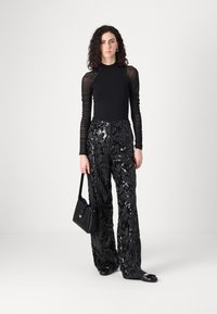 Top nero a maniche lunghe in tessuto trasparente, abbinato a pantaloni larghi neri con paillettes. La modella porta una piccola borsa nera e indossa delle ballerine nere.