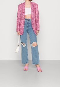 Blazer à motif floral rose, crop top blanc, jean large bleu clair déchiré, et sandales à talons roses avec brides croisées, accompagné d'un petit sac à main blanc.