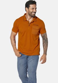 Orange polo shirt with a pocket, black stitching detail, short sleeves, and a fitted design, paired with blue jeans.