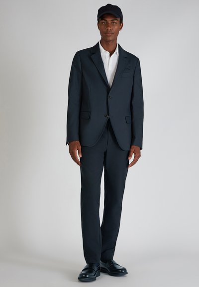 Marineblauer Anzug mit strukturiertem Blazer und gerade geschnittenen Hosen, kombiniert mit einem weißen Hemd und einer marineblauen Mütze. Aus glattem Stoff gefertigt und mit schwarzen Schuhen.