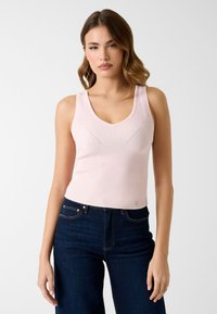 Débardeur rose pâle côtelé avec un col en V et une coupe ajustée, présentant des détails subtils sur la poitrine. Associé à un jean bleu foncé.