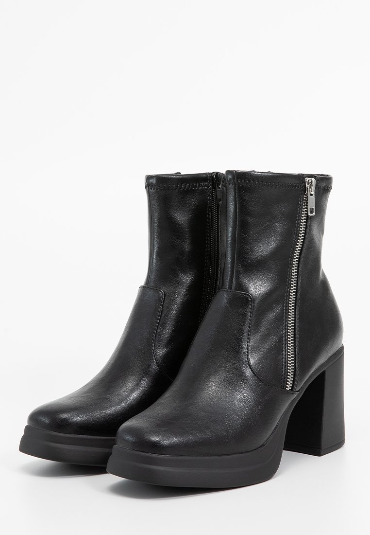 Bottines en cuir noir avec un talon épais, dotées de fermetures éclair argentées sur les côtés extérieurs et d'une texture lisse et brillante.