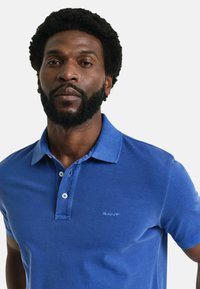 Sininen polo-paita, joka on valmistettu teksturoidusta kankaasta ja jossa on kolmipainikepanssari sekä lyhyet hihat. Paidassa on pieni brodeerattu logo rinnassa.