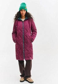 Prošívaný dlouhý kabát v tmavé burgundské barvě se zipem, kapucí a šňůrkami v tyrkysové barvě. Kombinován s hnědými kalhotami a světle hnědými botami. Měkká textura a teplo.