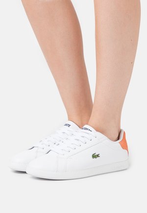 Baskets basses blanches avec logo crocodile vert et détail talon orange portées pieds nus sur fond blanc.