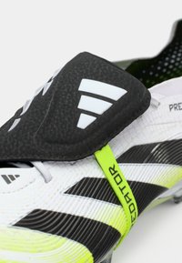 Close-up van een wit en zwart voetbalschoen met neongele accenten en een zwarte beschermflap over de veters met een wit logo.