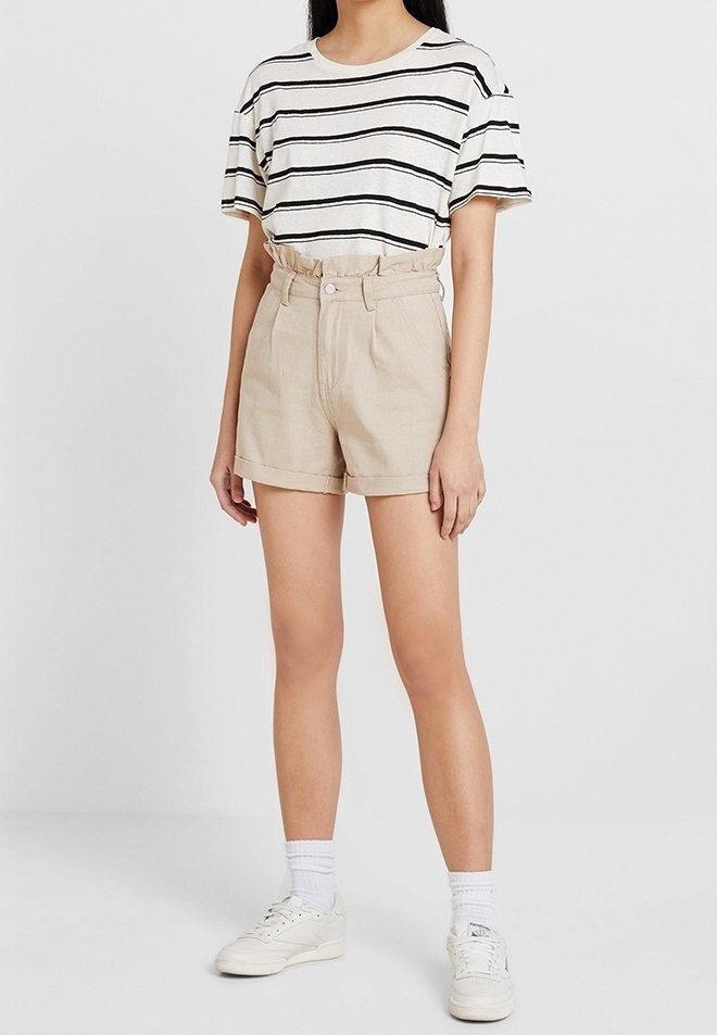 Femme portant un t-shirt à manches courtes rayé blanc et noir, un short beige taille haute, des chaussettes blanches et des baskets blanches, debout devant un fond uni.