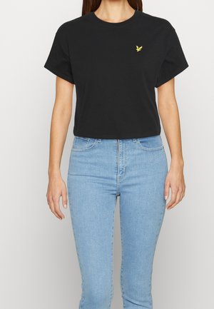 Frau trägt ein schlichtes schwarzes, kurzes T-Shirt mit kleinem gelbem Vogel-Logo auf der linken Brust und hellblaue Jeans mit hohem Bund.