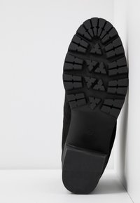 Semelle d'une botte noire avec des crampons profonds et un talon bloc, taille 40, appuyée contre un mur d'angle blanc et un sol.