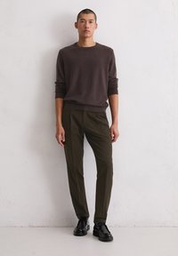 Junger Mann steht vor einer weißen Wand und trägt einen braunen Langarmpullover, olivgrüne Hose und schwarze Schnürschuhe.