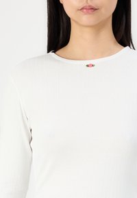 Vit ribbad topp med rundad halsringning och en liten broderad rosa rosdetalj nära kragen. Slät, åtsittande textur.