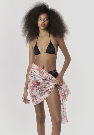 Mujer con cabello rizado y voluminoso, vestida con un bikini negro y un pareo floral, posando contra un fondo claro y liso.