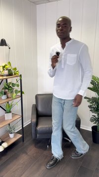 Camisa branca de manga longa com um bolso, jeans azul claro e sapatos pretos. Ambiente interior com plantas e uma cadeira preta em piso de madeira escura.