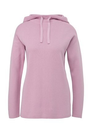 Femme portant un sweat à capuche rose clair avec cordons et un jean ajusté bleu foncé, debout devant un fond clair uni.