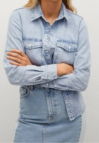 Chemise en denim bleu clair à manches longues, boutonnée sur le devant, avec deux poches poitrine et un col classique. Portée avec une jupe en denim bleu clair assortie.