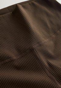 Hnedá rebrovaná látka s jemným leskom, s plochými švami a hladkou textúrou. Dizajn zahŕňa vysoký pás a priliehavý strih.