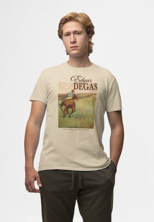 Junger Mann trägt ein beige T-Shirt mit Edgar Degas' Gemälde „Jockey in Blau auf einem kastanienbraunen Pferd“ und braunem Text über dem Kunstwerk.