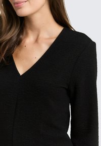 Schwarzes, strukturiertes Oberteil mit V-Ausschnitt. Aus ribbeligem Stoff mit subtiler Lichtreflexion, lange Ärmel und taillierte Silhouette.