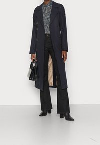 Marinblå trench coat med dubbelknäppning, stora knappar och en beige insida. Matchad med svarta utsvängda jeans och klackstövlar.