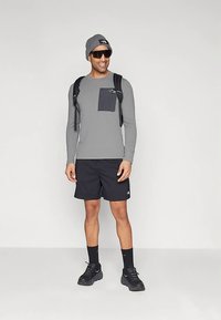 Férfi szürke hosszú ujjú pólóban, fekete rövidnadrágban, fekete tornacipőben, fekete zokniban, szürke sapkában, napszemüvegben és fekete hátizsákban áll fehér háttér előtt.