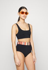 Fato de banho preto de duas peças com um top de decote redondo. A parte de baixo de cintura alta apresenta um cinto listrado multicolorido com detalhes em estampas de animais. Óculos de sol laranja.