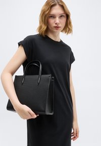 Νεαρή γυναίκα με κοντά ξανθά μαλλιά που φοράει μαύρο φόρεμα, κρατώντας μια μαύρη τσάντα με υφή και πάνω λαβές, πάνω σε απλό φόντο.