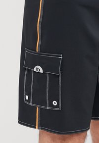 Shorts de bain noirs avec une rayure latérale orange, des coutures blanches et une poche à rabat avec un logo gris circulaire et une fermeture par bouton-pression.