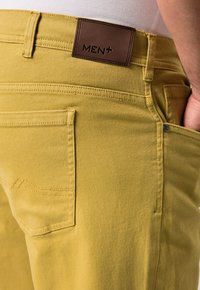 Žluté denimové džíny mají hnědou koženou nášivku s nápisem "MEN+" na pase. Obsahují standardní kapsy a viditelné švy.