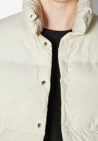 Ljus beige puffjacka med hög krage, som har tryckknappar och en veckad textur. Bärs över en mörk, ribbad tröja.