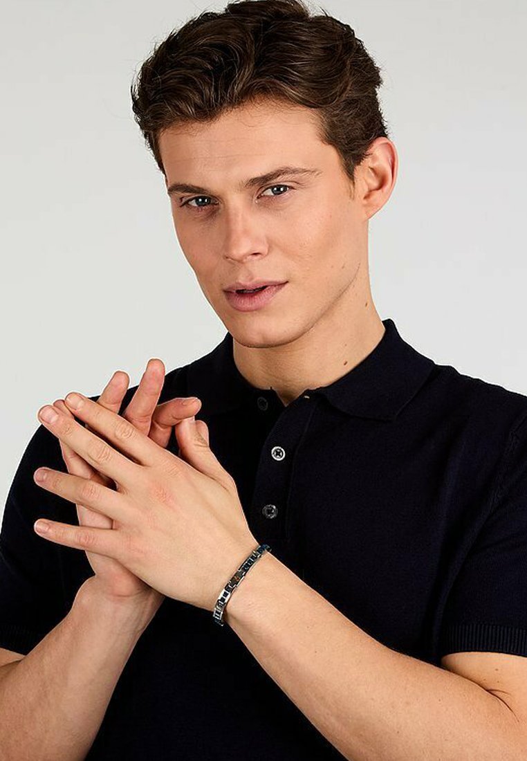 Metallarmband med rektangulära länkar i silver och svart, bärs på en hand. Personen har på sig en mörk polo t-shirt. Händerna är ihopknäppta.