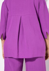Top viola, testurizzato, con maniche a tre quarti, caratterizzato da una piega sul retro e spacchi laterali. Abbinato a pantaloni coordinati, creando un look armonioso.