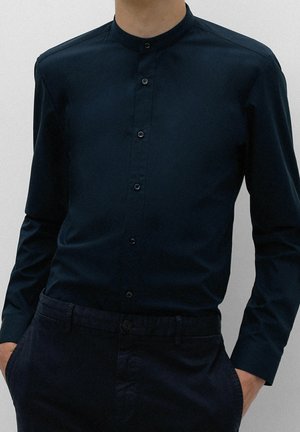 Mann trägt ein dunkelblaues Hemd mit Mandarinkragen und langen Ärmeln sowie passende dunkle Hose, Hände in den Taschen.