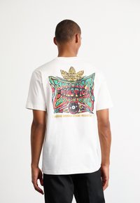 Vit bomullströja med en färgglad grafisk design på baksidan som har en ögonmotiv och texten "Adidas Originals Studio Production".