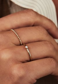 Bague en or avec une fine bande et un petit accent de diamant rond. La main porte également un bracelet en or avec des perles. Texture de peau lisse visible.