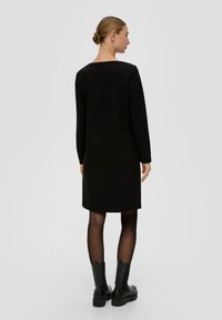 Czarna, długa sukienka z długim rękawem, okrągłym dekoltem, o teksturowanej tkaninie, do kolan, noszona z czarnymi botkami za kostkę i przezroczystymi czarnymi rajstopami.