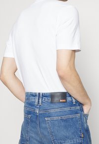 Valkoinen puuvillapaita lyhyillä hihoilla, yhdistettynä korkeavyötäröisiin sinisiin farkkushortseihin, joiden vyötäröllä on musta nahkapäällyste, johon on kirjailtu "BOSS".