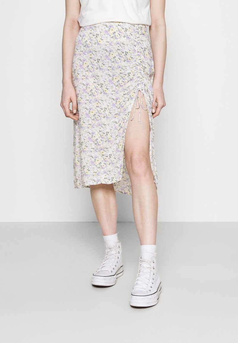 Blomstret midi-nederdel i lyse lilla med gule accenter, med detalje af siderynker og justerbare bånd. Sammen med hvide high-top sneakers.