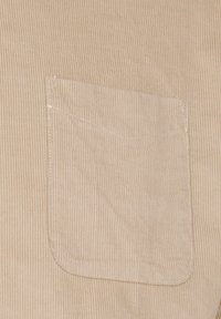 Tejido de sarga beige con un solo bolsillo en el pecho de borde redondeado y costura blanca, que muestra una textura acanalada vertical.
