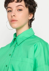 Camisa de manga curta em verde brilhante, feita de um tecido leve. Apresenta um colarinho com botões e um bolso no peito. Textura suave.