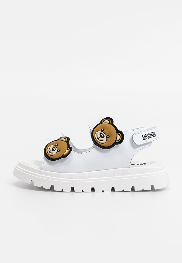 Sandales blanches avec une semelle épaisse blanche, ornées de deux appliqués en forme de tête d'ours brun. Fabriquées en matériau lisse avec un détail de bande réglable.