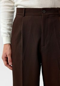 Bruine geribde broek met een getailleerde pasvorm, voorzien van een gladde textuur en een knoopsluiting bij de taille. Een wit gebreid topje is zichtbaar.