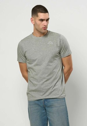 Mężczyzna z krótkimi ciemnymi włosami, ubrany w jasnoszary t-shirt z małym białym logo na piersi i jasnoniebieskie dżinsy, stojący przy białej ścianie.