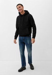 Czarna bluza z kapturem z przednią kieszenią, zestawiona z niebieskimi dżinsami. Model ma na sobie czarne botki. Tło jest jednolicie białe.