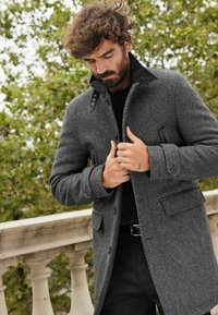 Grå tekstureret frakke med krave, sorte accenter og knapdetaljer. Parret med en sort sweater, båret over sorte bukser. Dekorative knapper synlige.