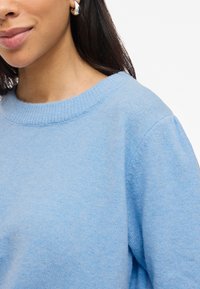 Sweater de malha azul claro, com decote redondo, apresentando uma textura suave e ombros caídos. Detalhe de destaque no decote.