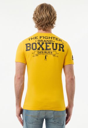Uomo con capelli biondi mossi che indossa una maglietta gialla con testo nero "THE FIGHTER BRAND BOXEUR DES RUES" e una piccola silhouette di pugile sulla schiena.