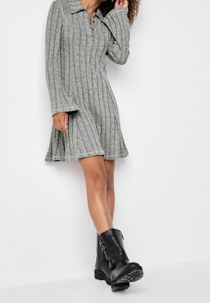 Femme portant une robe tricotée grise à côtes avec des manches évasées et des bottines noires à boucles, debout contre un mur blanc.