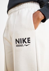 Világosszürke melegítőnadrág texturált felülettel, fekete "NIKE" logóval és praktikus oldalsó zsebbel. Rugalmas derékpánt található.