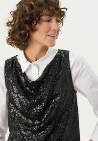 Sort pailletetop med draperet design, der har en rund halsudskæring og ærmeløs skæring, lagd over en hvid skjorte med knapper.