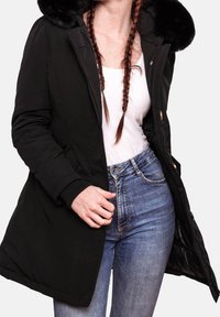 Manteau d'hiver matelassé noir avec capuche bordée de fourrure, doté d'une fermeture éclair, de poches latérales et de bordures côtelées. Porté sur un haut blanc et un jean.